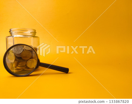 The image displays a clear glass jar filled with coins and a magnifying glass resting against it, set against a yellow background. The image displays a clear glass jar filled with coins and a magnifying glass resting against it, set against a yellow background. 133867820