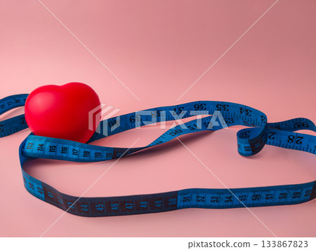 The image features a red heart object encircled by a blue tape measure, set against a pink background. 133867823