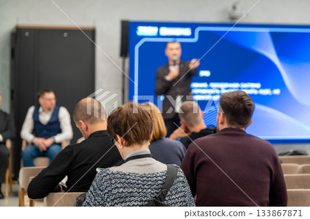 Conference presentation in modern hall with speaker on stage and attentive audience Conference presentation in modern hall with speaker on stage and attentive audience 133867871