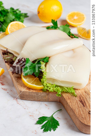 Fresh squid prepared for cooking, surrounded by herbs and lemon, placed on a wooden chopping board in a bright kitchen setting Fresh squid prepared for cooking, surrounded by herbs and lemon, placed on a wooden chopping board in a bright kitchen setting 133867976