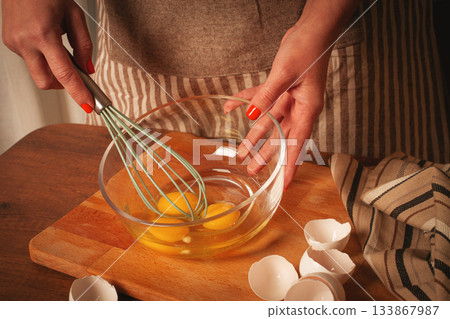Whisking fresh eggs in a clear bowl on a wooden countertop during morning cooking session 133867987