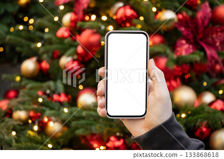 Smartphone with empty blank screen in mans hand against Christmas tree with light garland. Screen display for Festive Xmas Holiday Background. Space for text. Christmas advertising, smartphone app Smartphone with empty blank screen in mans hand against Christmas tree with light garland. Screen display for Festive Xmas Holiday Background. Space for text. Christmas advertising, smartphone app 133868618