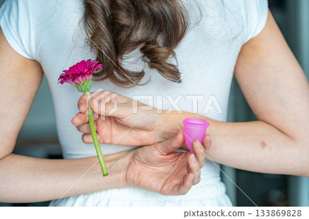 Woman holding a pink menstrual cup and a flower behind her back. Concept of feminine health, natural care, and sustainable period products with symbolic floral reference. Woman holding a pink menstrual cup and a flower behind her back. Concept of feminine health, natural care, and sustainable period products with symbolic floral reference. 133869828