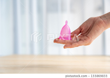 Close-up of a pink menstrual cup standing upright on a woman open palm over wooden table. Feminine hygiene product promoting zero waste, comfort, and sustainable period care. 133869833