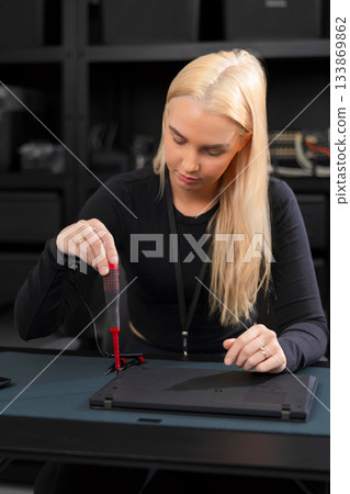 Young Caucasian woman with blonde hair repairs laptop using a screwdriver in a tech workshop 133869862