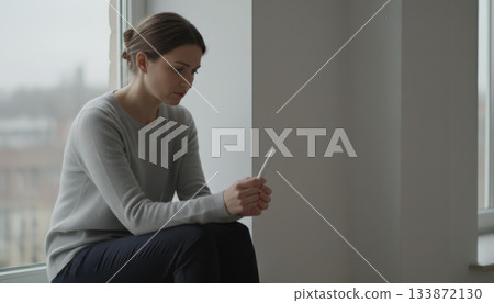 Woman sitting by a window, holding a pregnancy test stick, looking dejected, experiencing anxiety and stress while contemplating infertility issues and the desire for a family 133872130