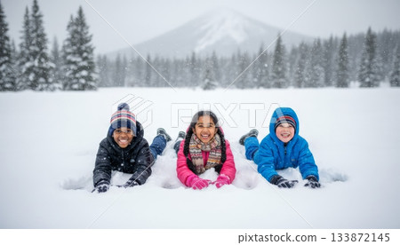 Three happy children of different ethnicities are playing in the snow, smiling and looking at the camera, enjoying outdoor winter activities in a snowy forest landscape with mountains 133872145