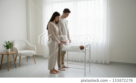 Loving parents standing beside a hospital bassinet, looking down at their sleeping newborn baby, sharing a tender and intimate moment after birth in a bright and minimalist room 133872276