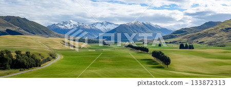 Stunning panorama of new zealand's countryside features lush green meadows, a winding road, rows of trees, and majestic snow capped mountains under a cloudy sky 133872313