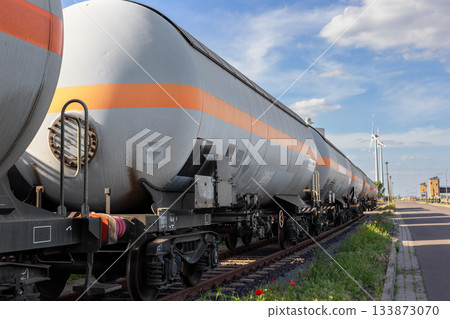 Long row of railroad tank railcars stands on sunlit tracks blue sky background. Industrial scene metal surfaces, wheels wind turbines. Energy commodity industry global trade logistics business Long row of railroad tank railcars stands on sunlit tracks blue sky background. Industrial scene metal surfaces, wheels wind turbines. Energy commodity industry global trade logistics business 133873070