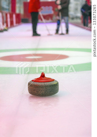 Single red curling stone on ice with blurred players in background during match. Friendly game of curling, teamwork, sport strategy, decision moment, winter active lifestyle 133873265