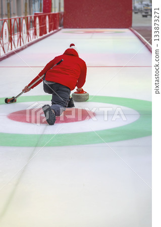 Outdoor curling player delivering stone towards circular target on frozen rink. Concept of winter sport precision, control and focused movement 133873271