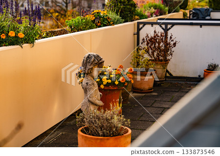 Terrace Garden With Flower Pots And Stone Statue: Sunlit Outdoor Arrangement Of Colorful Blossoms And Decorative Sculpture In Warm Autumn Atmosphere 133873546