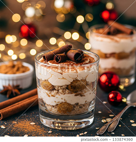 A festive layered dessert in a glass, adorned with chocolate curls, sits amidst holiday decorations and warm glowing lights, inviting celebration 133874360