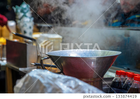 A metal pot and fryer setup releasing steam in the outdoor kitchen at a christmas market food stall, with bottles and wrapped supplies arranged on the counter. 133874419