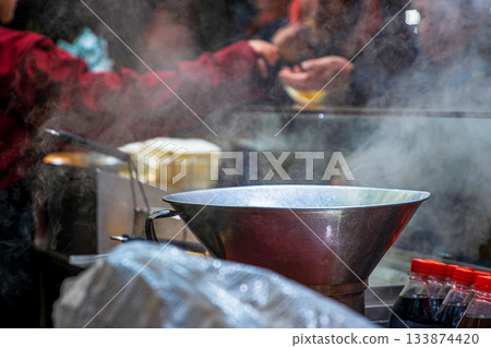 A steaming metal pot at a christmas market food stall with vendors serving customers in the background, surrounded by bottles and wrapped supplies. 133874420