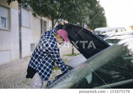 Young man diagnosing car engine on residential street during daytime Young man diagnosing car engine on residential street during daytime 133875576