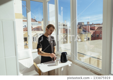 Young individual pours steaming water into cup at daylight window Young individual pours steaming water into cup at daylight window 133875919