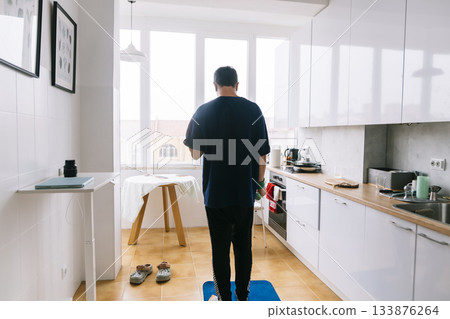 person monitoring body weight in bright kitchen, man using scale for fitness tracking near window light 133876264