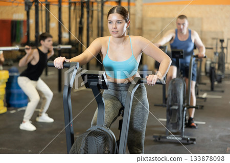 Young dedicated active strong fit woman doing cardio workout on stationary bicycle as hard cross-training in gym indoors 133878098