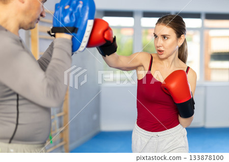 Athletic woman learns to box - hitting punch mitts. Woman with her trainer at gym Athletic woman learns to box - hitting punch mitts. Woman with her trainer at gym 133878100
