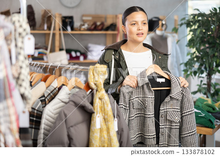 Interested asian woman standing in clothing shop and looking new coat for autumn-winter season. Shopping and seasonal sales concept 133878102