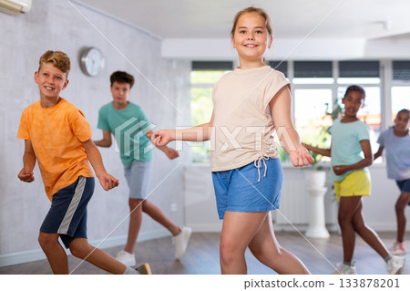Positive juvenile girl engaged in Rock 'n' Roll dance in training room with children's group Positive juvenile girl engaged in Rock 'n' Roll dance in training room with children's group 133878201