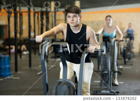 Motivated young man warming up and pedaling on exercise bike during group CrossFit workout in gym health center 133878202
