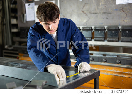 Guy working at metallurgical plant carefully measures sheet of iron using tape measure 133878205