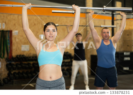 Energetic young female athlete practicing sit-ups with rod near other people gym Energetic young female athlete practicing sit-ups with rod near other people gym 133878231