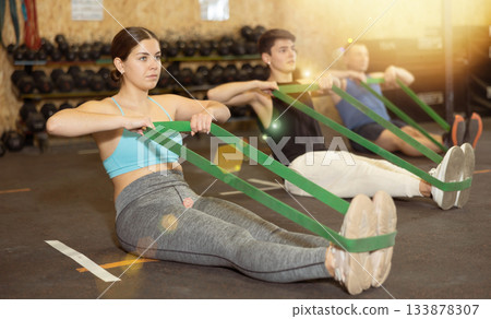 Motivated young woman practicing exercises with stretch rope sitting near other people in gym Motivated young woman practicing exercises with stretch rope sitting near other people in gym 133878307