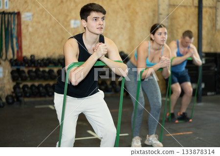 Active young man wearing sport clothes using resistance band during group workout in gym. Functional training and CrossFit concept. Active young man wearing sport clothes using resistance band during group workout in gym. Functional training and CrossFit concept. 133878314