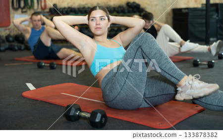 Concentrated young fitness woman doing abdominal exercise on yoga mat during group workout in gym. CrossFit healthy concept. 133878382