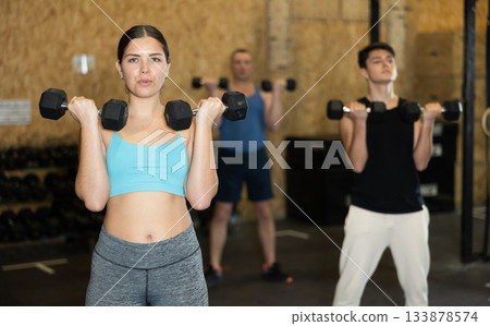 Young dedicated active strong fit woman doing exercises for shoulders while pushing up dumbbells in CrossFit center 133878574