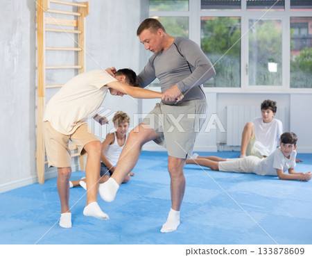 Male karate instructor training little child in dojo or jiu-jitsu in gym. Children sitting on floor around coach 133878609
