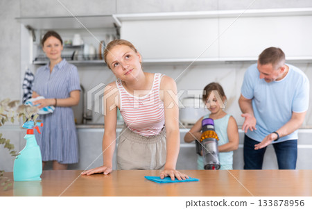 Teen girl cleans table with rag, family clean kitchen in background 133878956