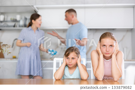 Frightened and frowning children sit at kitchen table during mom and dad quarrel. 133879082