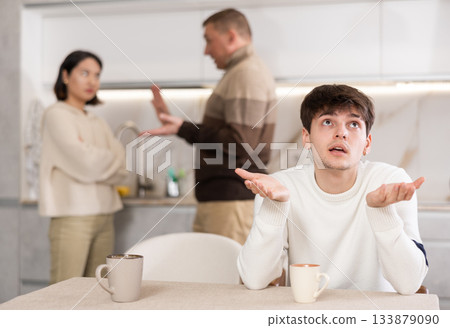 Angry young guy is tired of domestic quarrels. Relatives quarreling in background Angry young guy is tired of domestic quarrels. Relatives quarreling in background 133879090