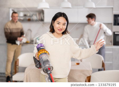 Girl vacuuming floor while his relatives help tidy up room 133879195