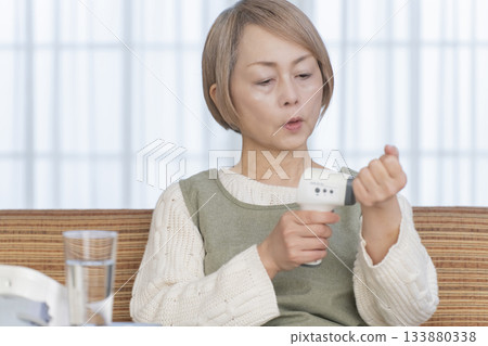 Senior woman measuring her temperature with a thermometer in the living room Senior woman measuring her temperature with a thermometer in the living room 133880338