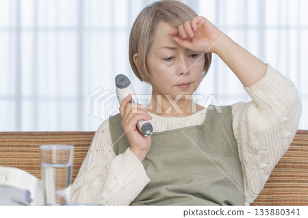Senior woman measuring her temperature with a thermometer in the living room Senior woman measuring her temperature with a thermometer in the living room 133880341
