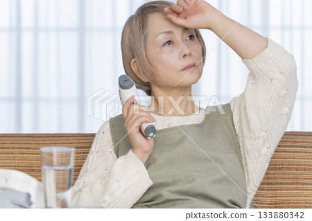Senior woman measuring her temperature with a thermometer in the living room 133880342