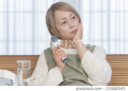 Senior woman measuring her temperature with a thermometer in the living room Senior woman measuring her temperature with a thermometer in the living room 133880345