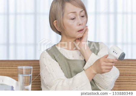 Senior woman measuring her temperature with a thermometer in the living room Senior woman measuring her temperature with a thermometer in the living room 133880349