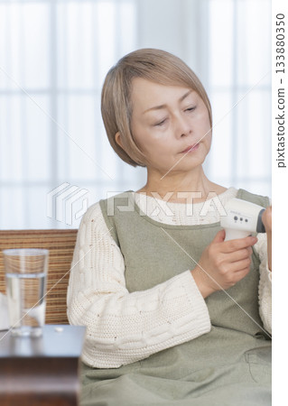 Senior woman measuring her temperature with a thermometer in the living room Senior woman measuring her temperature with a thermometer in the living room 133880350