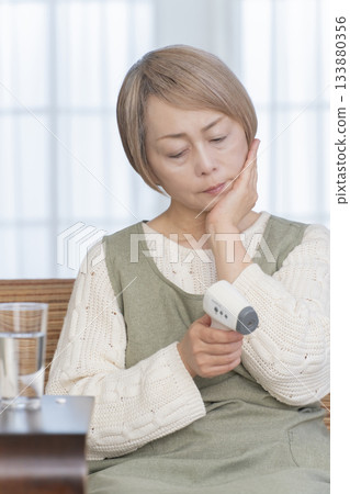 Senior woman measuring her temperature with a thermometer in the living room Senior woman measuring her temperature with a thermometer in the living room 133880356
