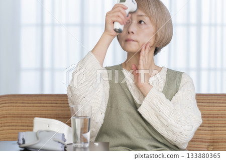 Senior woman measuring her temperature with a thermometer in the living room 133880365