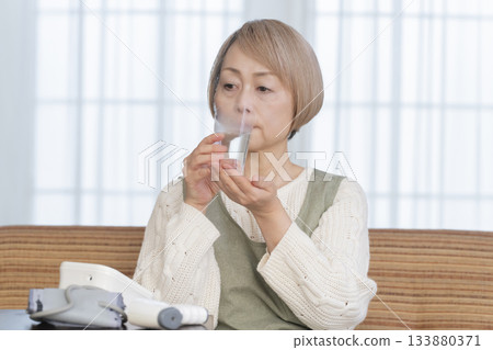 Senior woman measuring her temperature with a thermometer in the living room Senior woman measuring her temperature with a thermometer in the living room 133880371