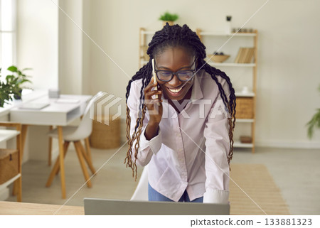 Happy young African American businesswoman using laptop and talking on mobile phone Happy young African American businesswoman using laptop and talking on mobile phone 133881323