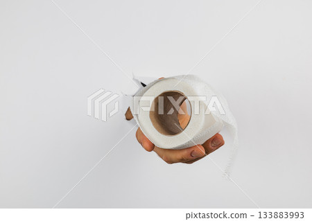 Hand holding a roll of toilet paper with torn wrapping in bright indoor light Hand holding a roll of toilet paper with torn wrapping in bright indoor light 133883993